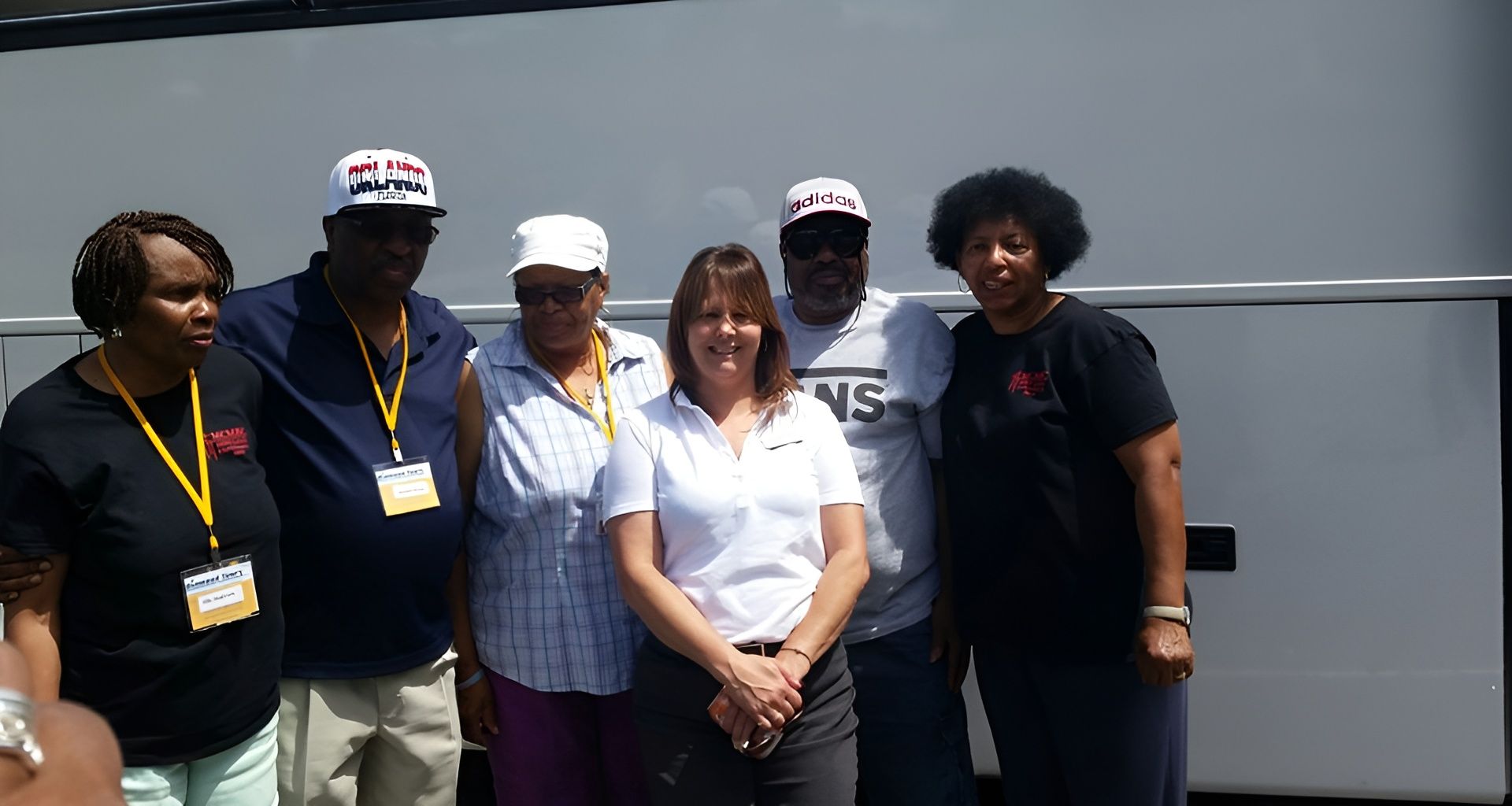 Group of people posing by a bus. Several wear hats. Woman in center wears a logo shirt. Outdoors, sunny.