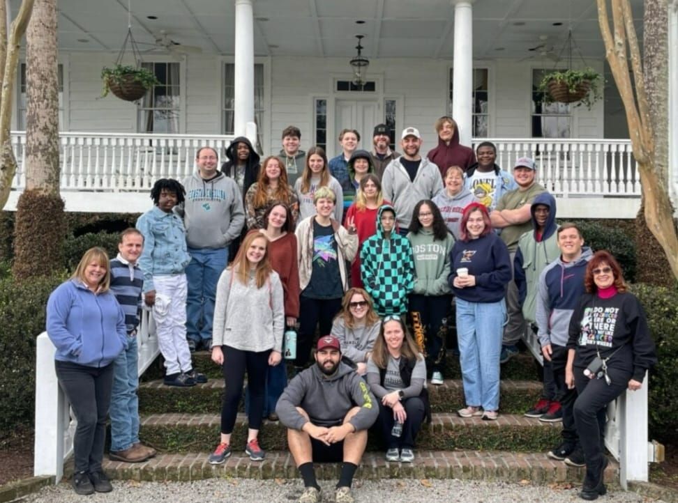 Group of people standing on steps in front of a white house with a porch.