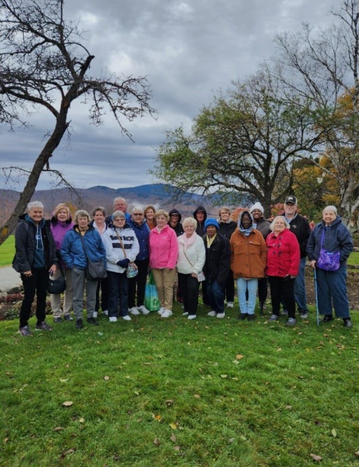 Group of people standing on green grass, possibly a tour group, with mountains in the background. Overcast day.