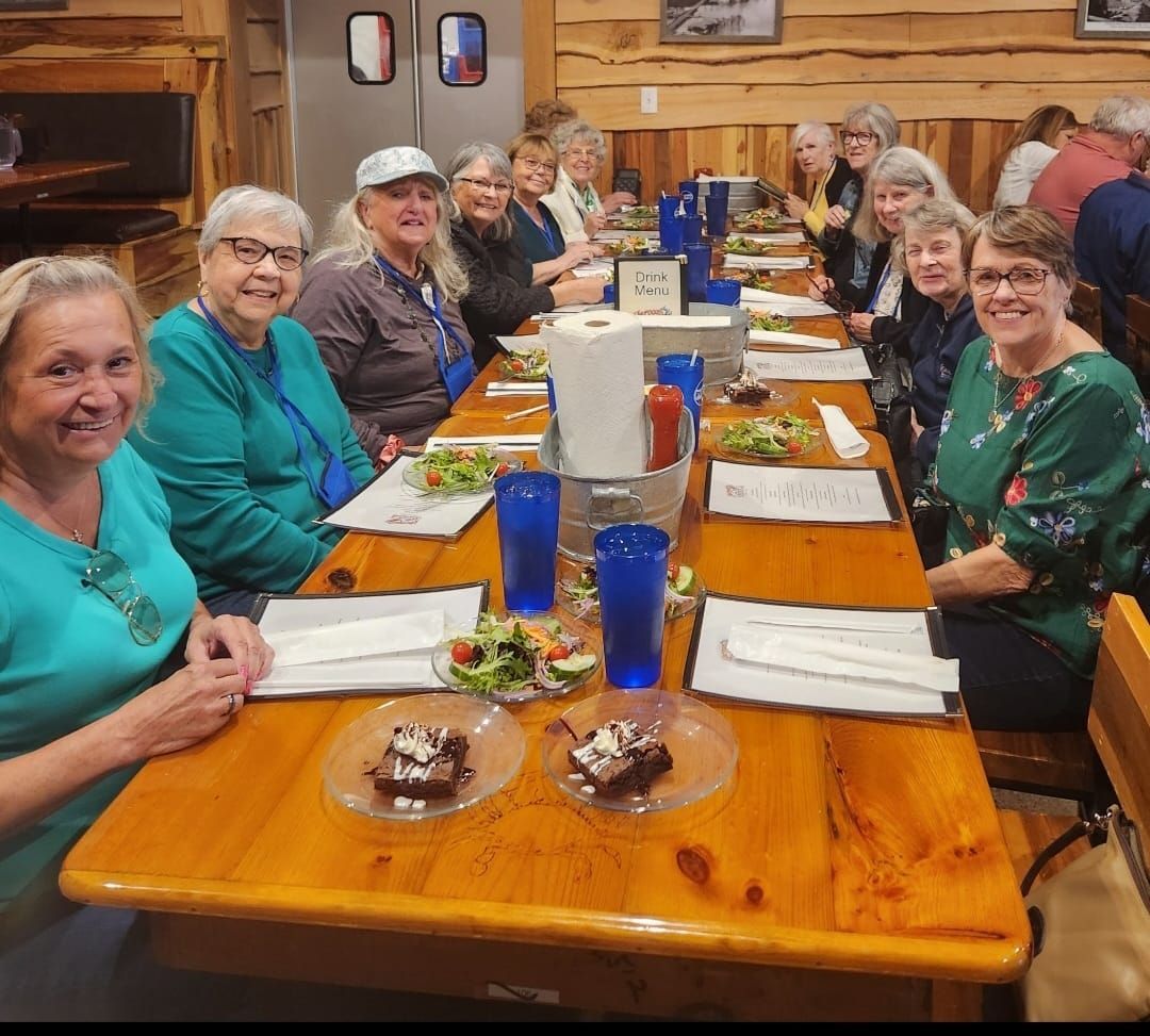 A group of people seated around a long wooden table, some with food, inside a restaurant.