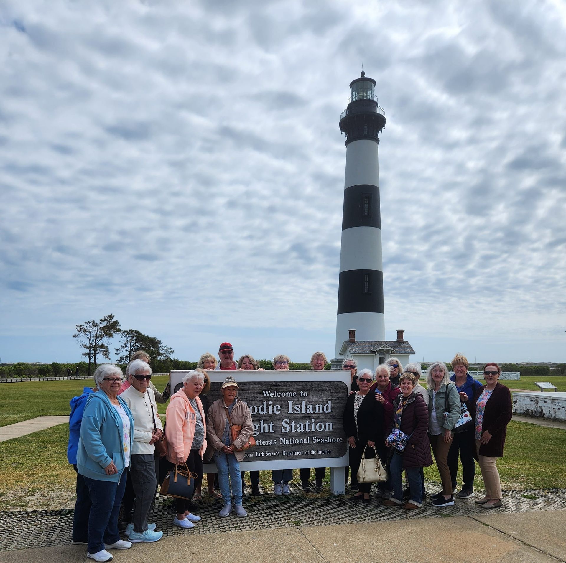 Group of people poses with a sign near a black and white lighthouse under a cloudy sky.