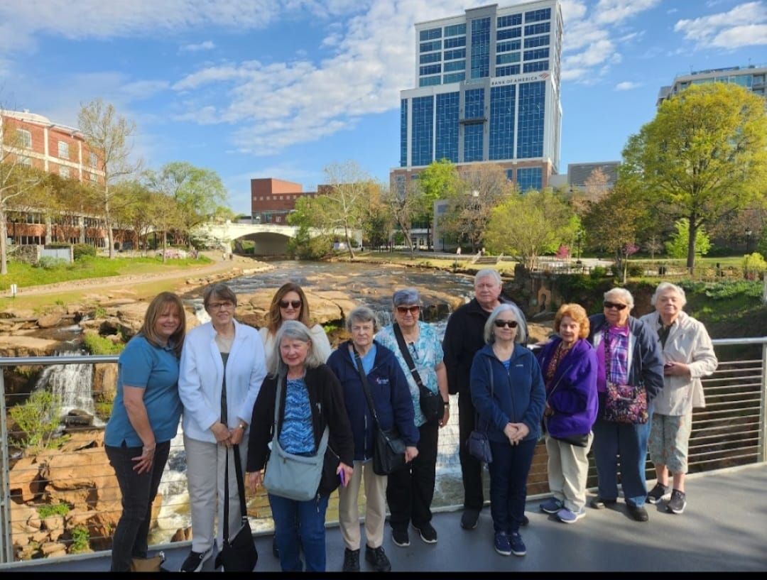 Group of people posing by a river and city buildings under a blue sky with clouds.