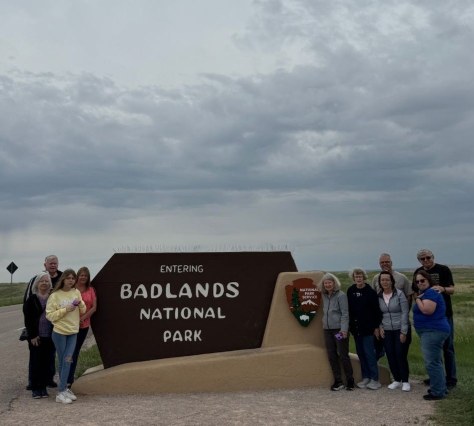 Group poses at Badlands National Park sign under a cloudy sky.