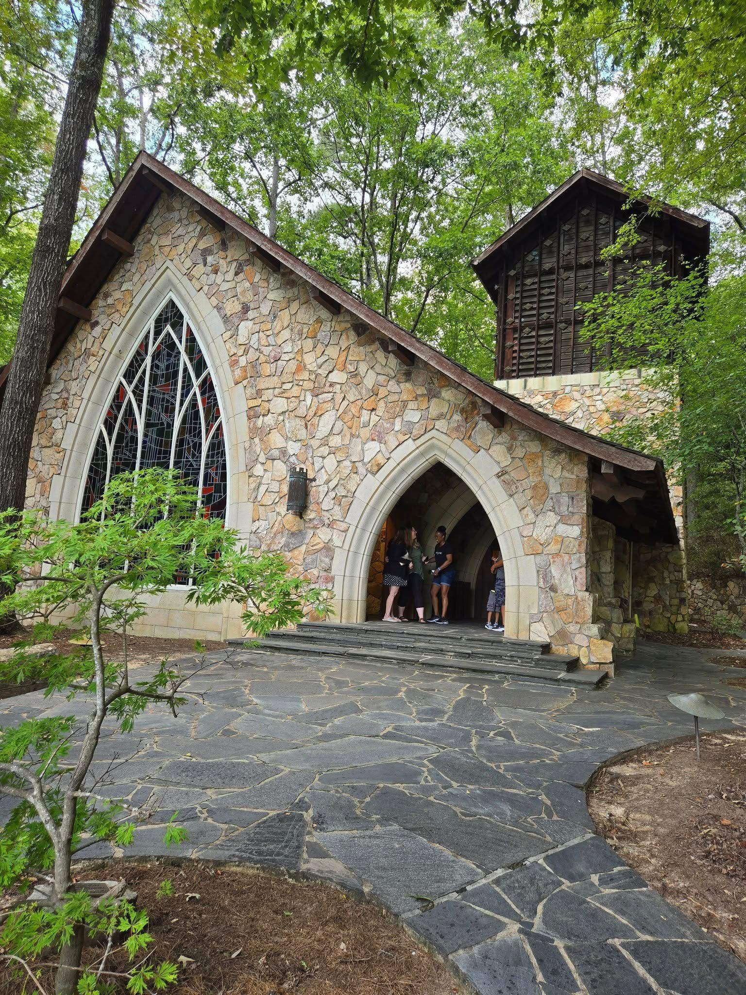 Stone chapel with arched entrance, stained glass, and a small tower, set in a wooded area.
