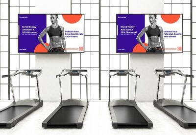 Loopbanden en digitale schermen in een sportschool. Op de schermen worden fitnessdiensten geadverteerd met een vrouw die aan het sporten is.