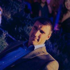 A man with short dark hair in a blue button-down shirt and brown jacket, smiling slightly in a dimly lit, blurred setting.