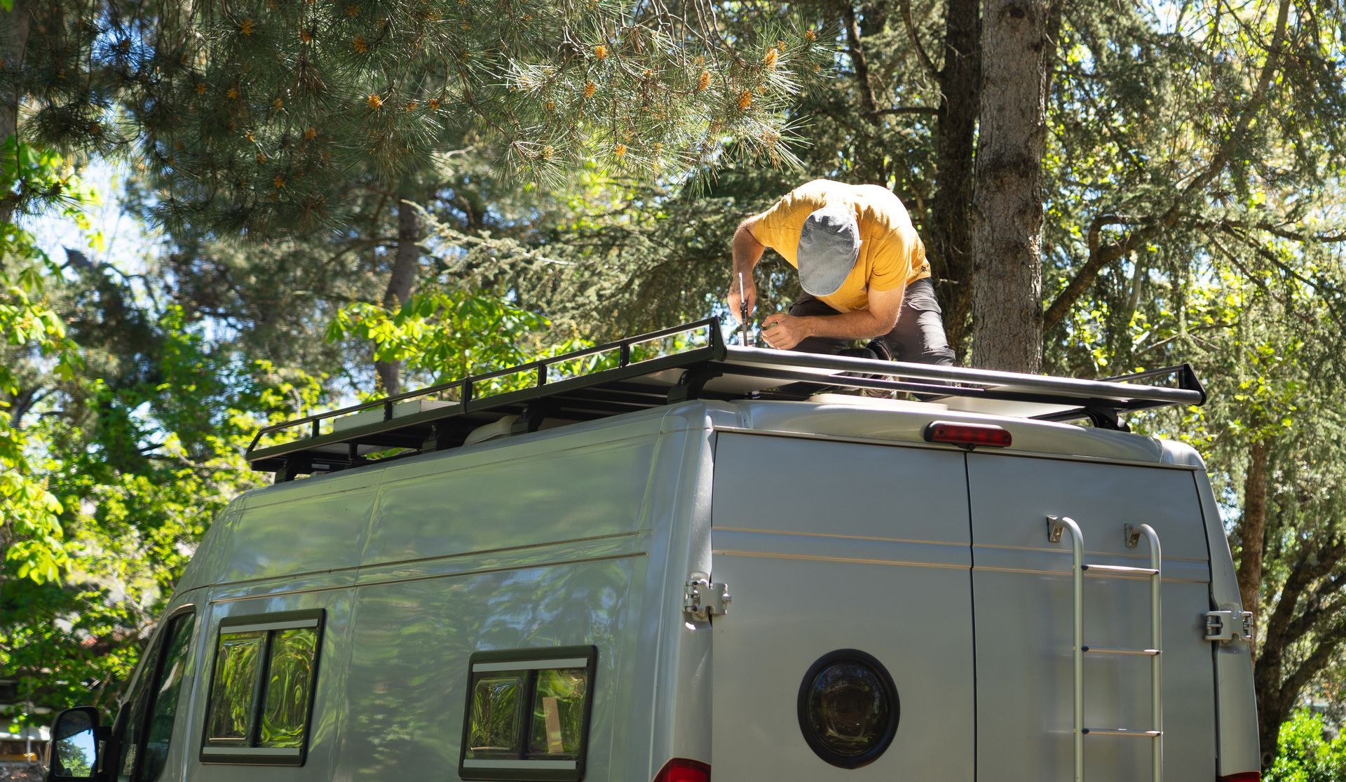 Person on roof of a white van in a wooded area, adjusting a roof rack.