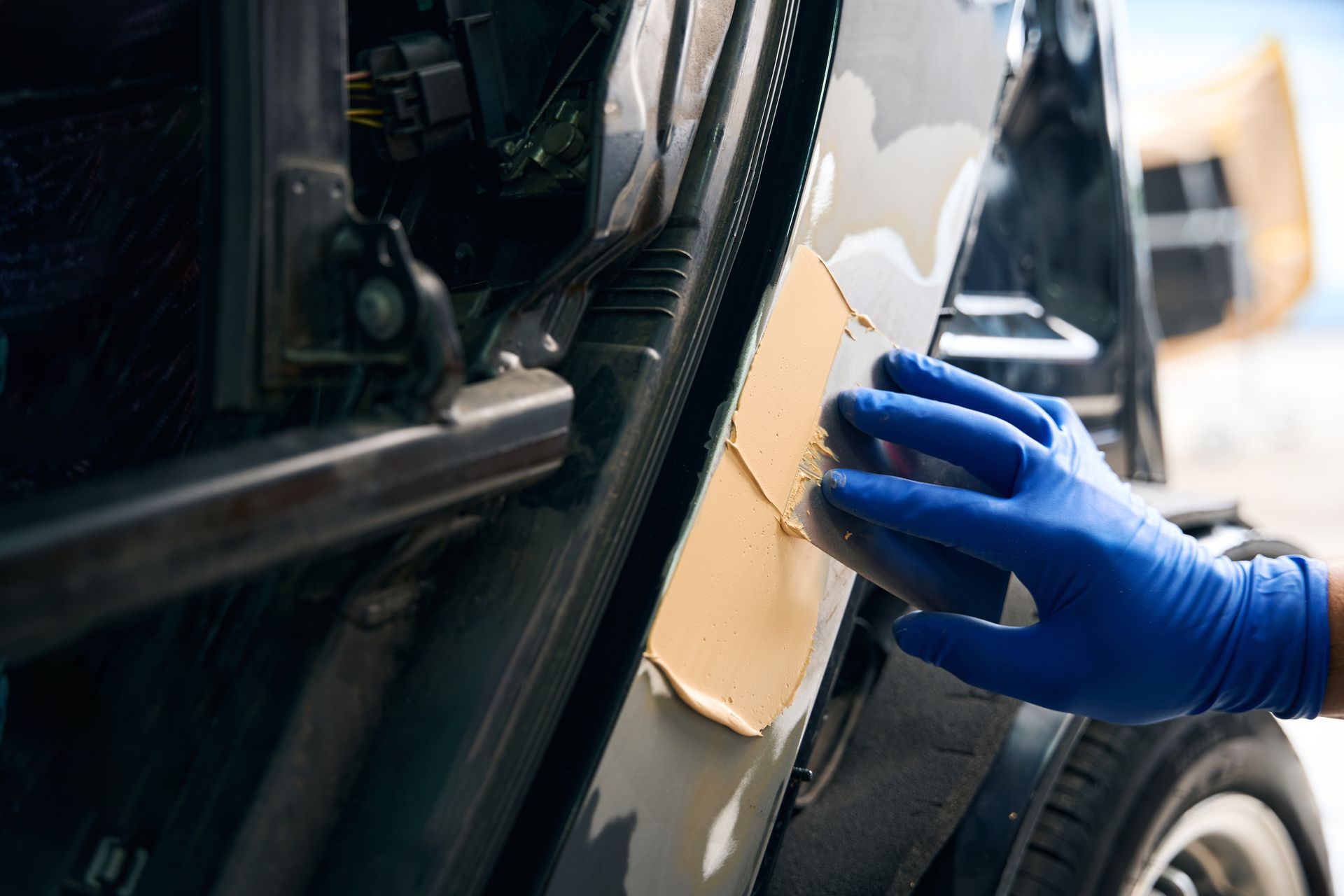 Blue-gloved hand applying body filler to a car door panel in a workshop