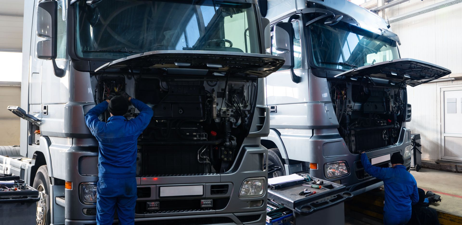 Two mechanics inspecting silver trucks in a repair garage with hoods open