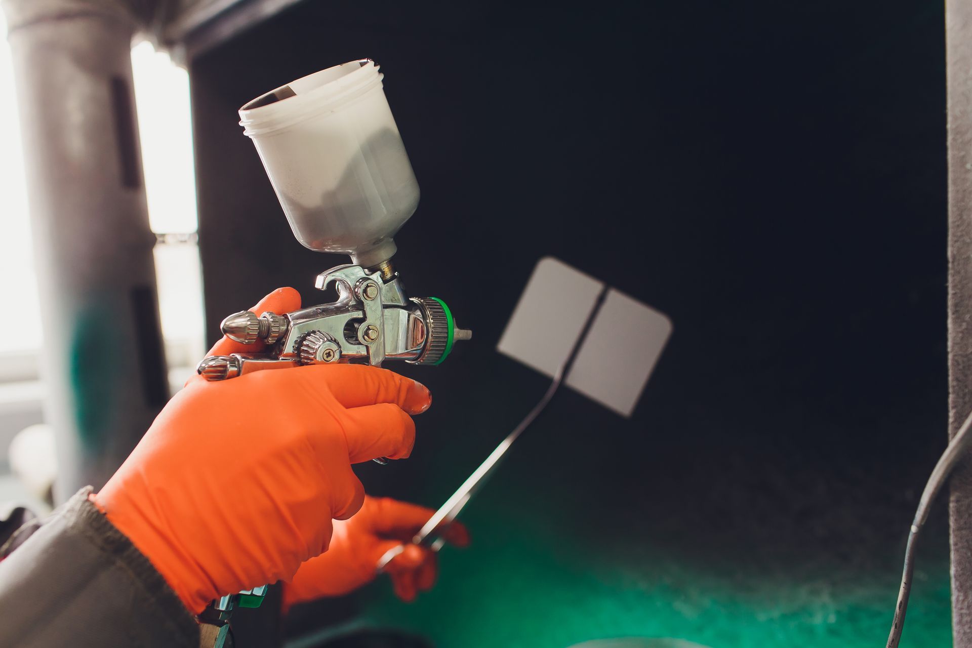 Orange-gloved hands spray-painting a small white object with a paint gun in a dark workshop