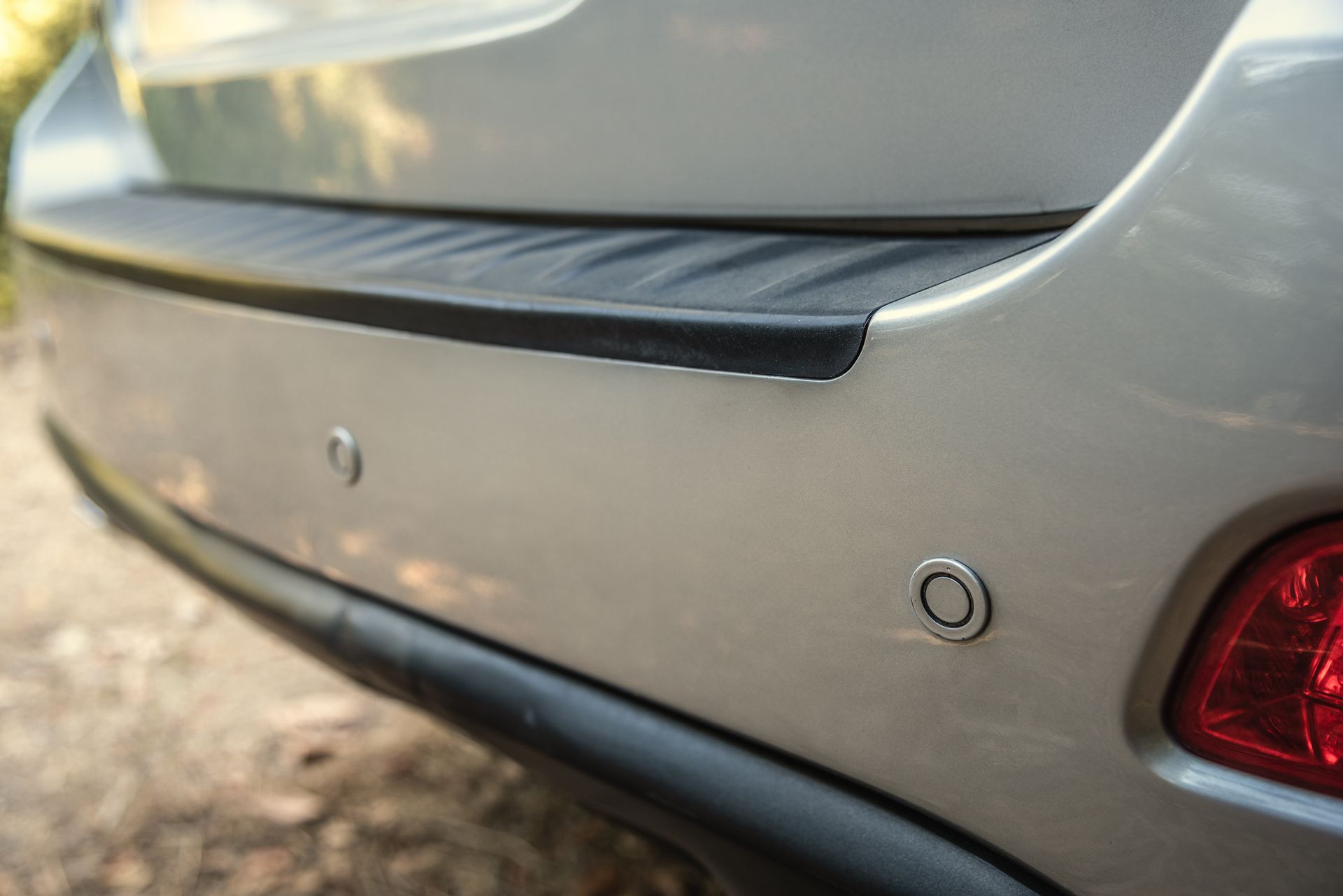 Close-up of a silver car’s rear bumper with parking sensors and a red taillight.