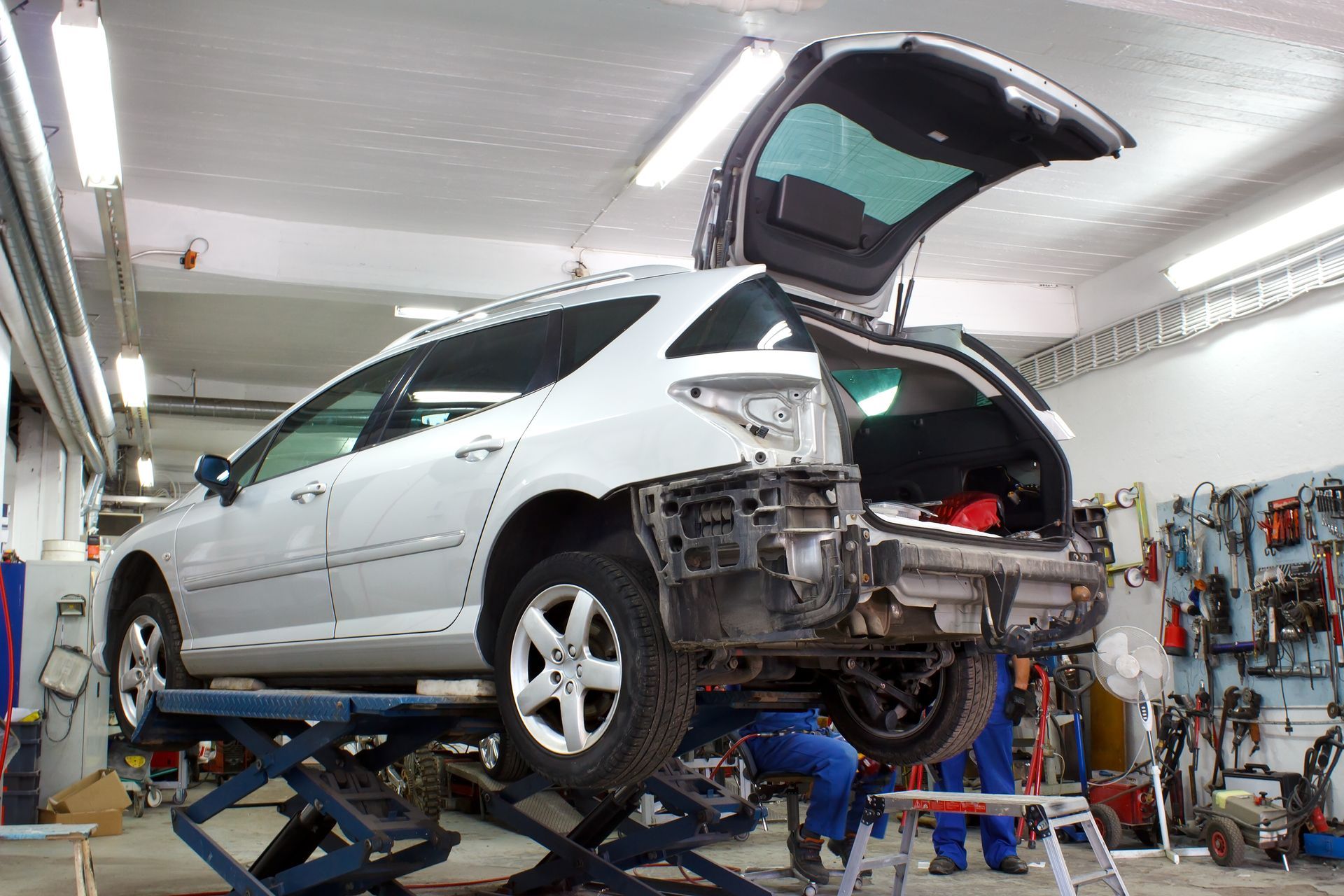 Silver car on a lift in an auto repair shop with its rear bumper removed and trunk open