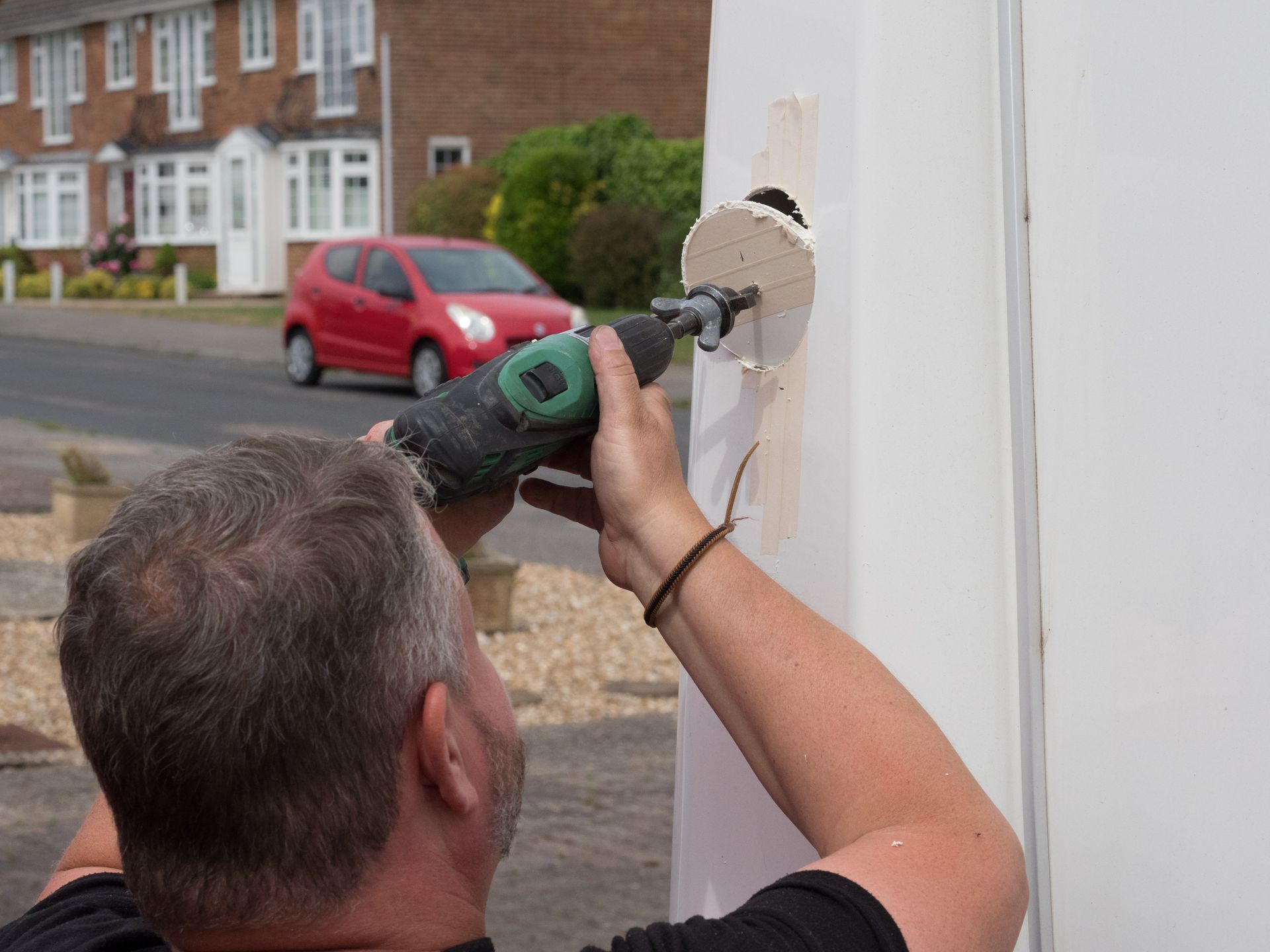 Person using a power drill to install or remove a white wall fixture outdoors near parked cars