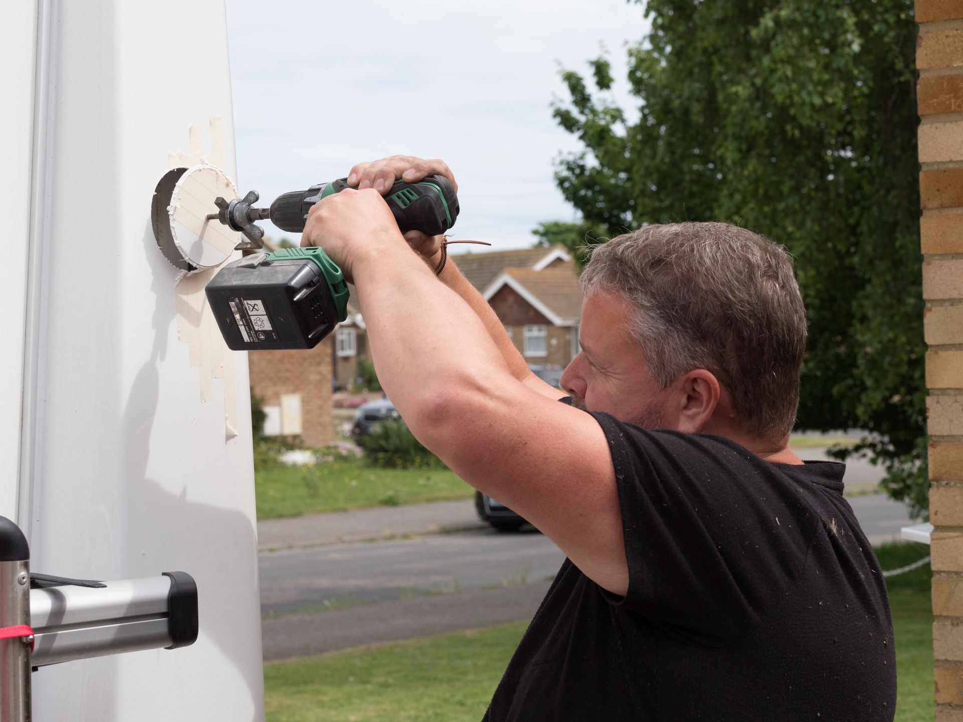 Man drilling into a white wall beside a brick house, outdoors in daylight.