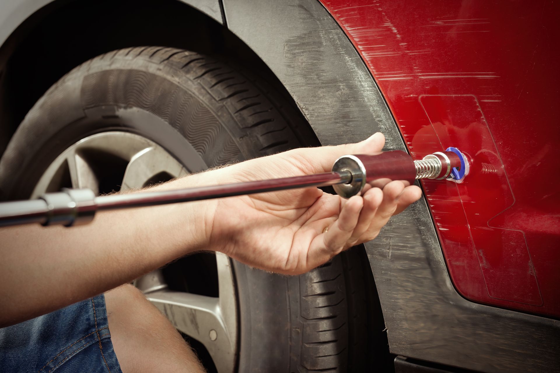 Person using a long tool to repair a red car’s body near the wheel.