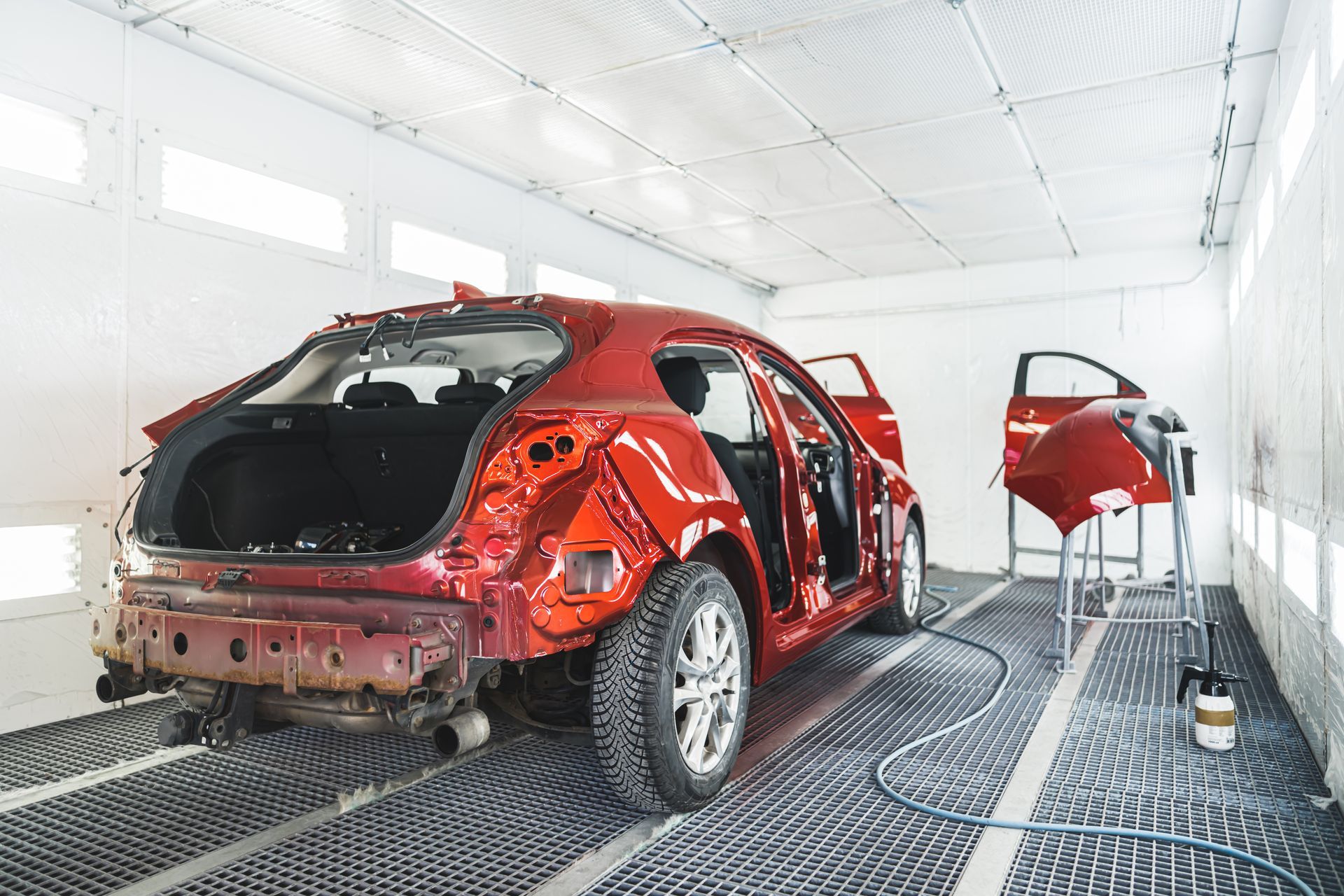 Red car body in a paint booth, stripped and undergoing auto repair with open doors and a detached panel.