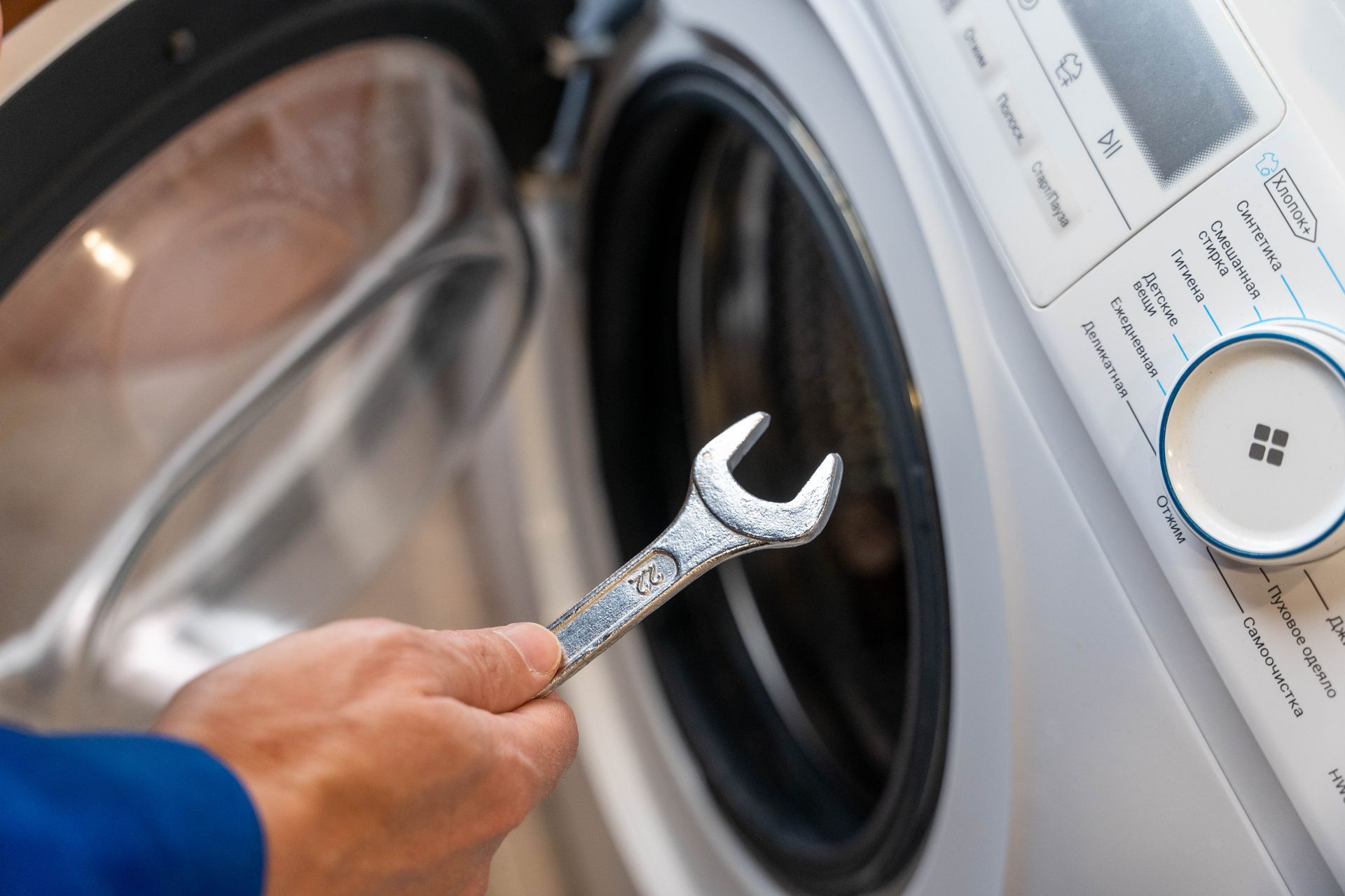 Hand holding a wrench near the open door of a white front-loading washing machine for repair