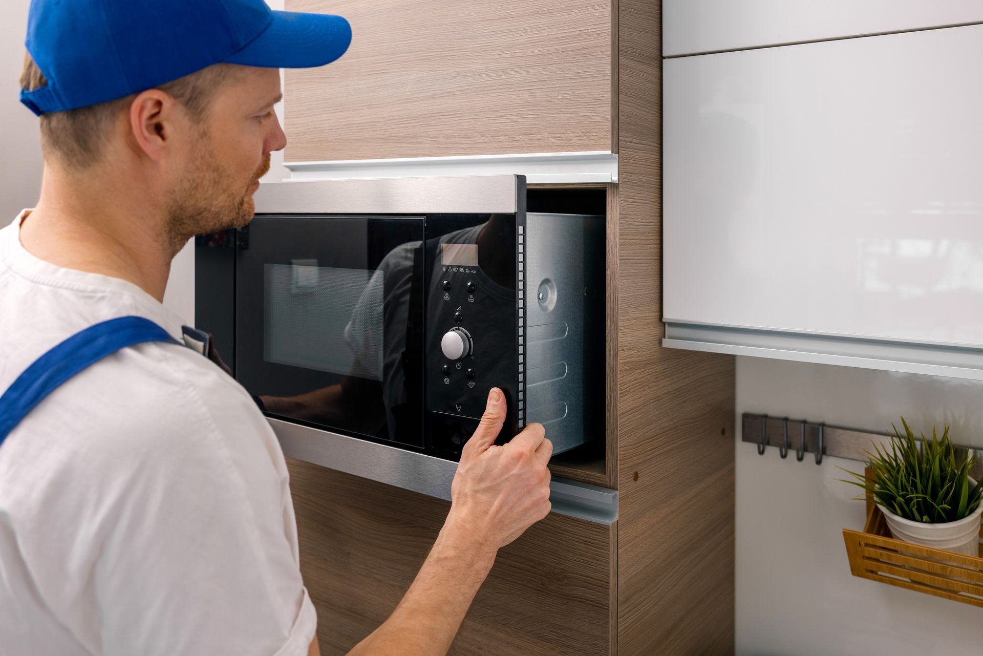 Worker opening a built-in microwave in a modern kitchen, with a white cabinet and small plant nearby.