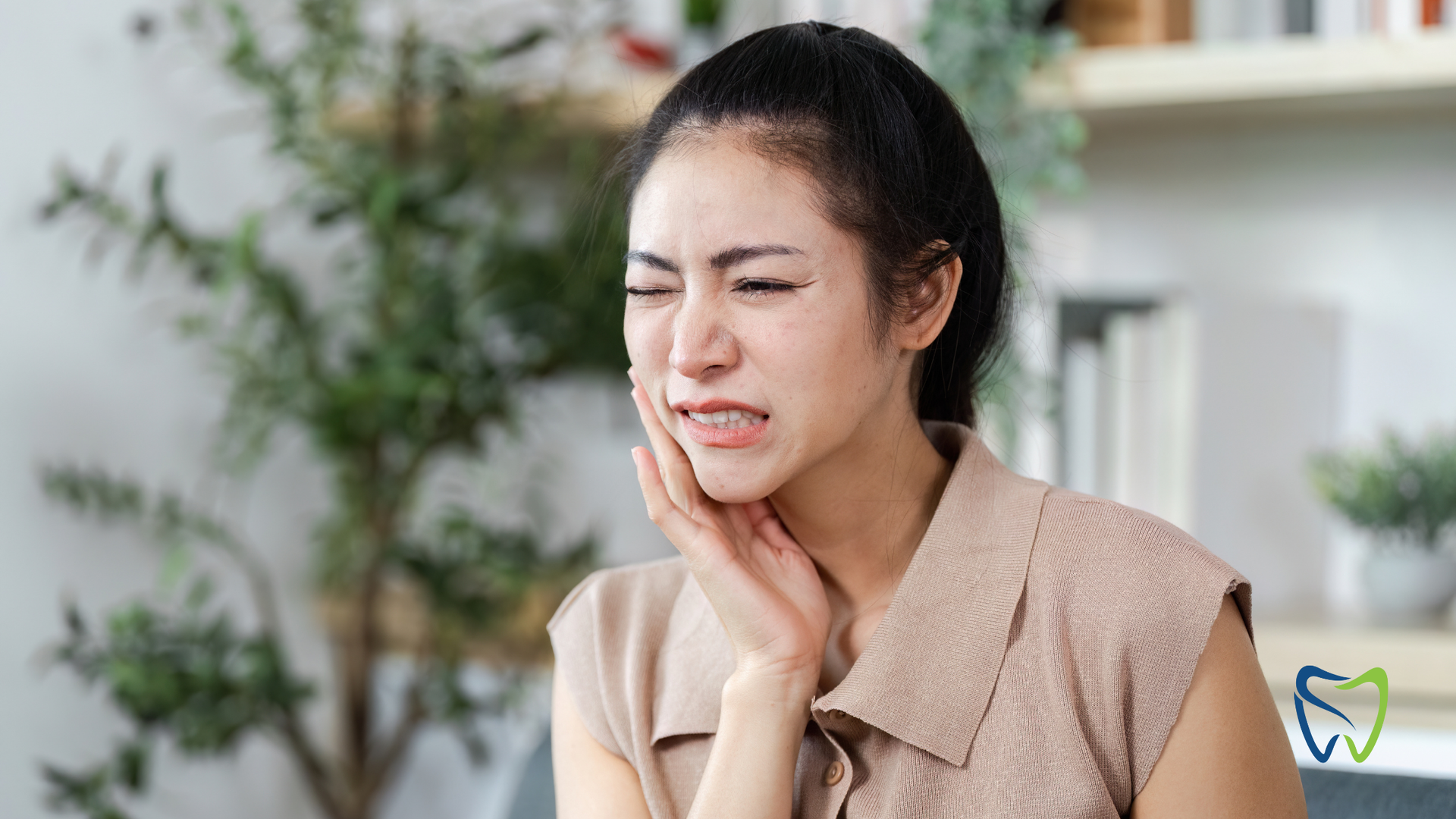 Woman with pained expression, holding cheek, likely experiencing toothache; indoor setting.