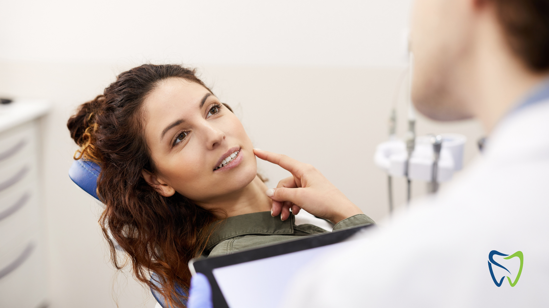 Woman in dental chair, looking at dentist, pointing to her jaw. Clinic setting.