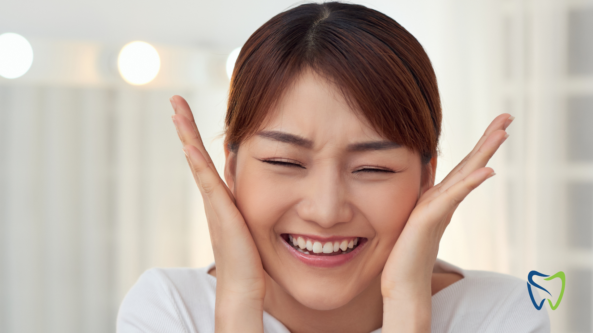 Woman with closed eyes smiling, hands on cheeks, in front of soft, bright lights.