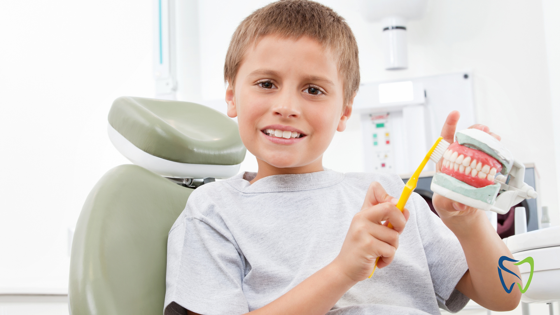 Boy in dentist's chair smiles, holding toothbrush and model teeth.