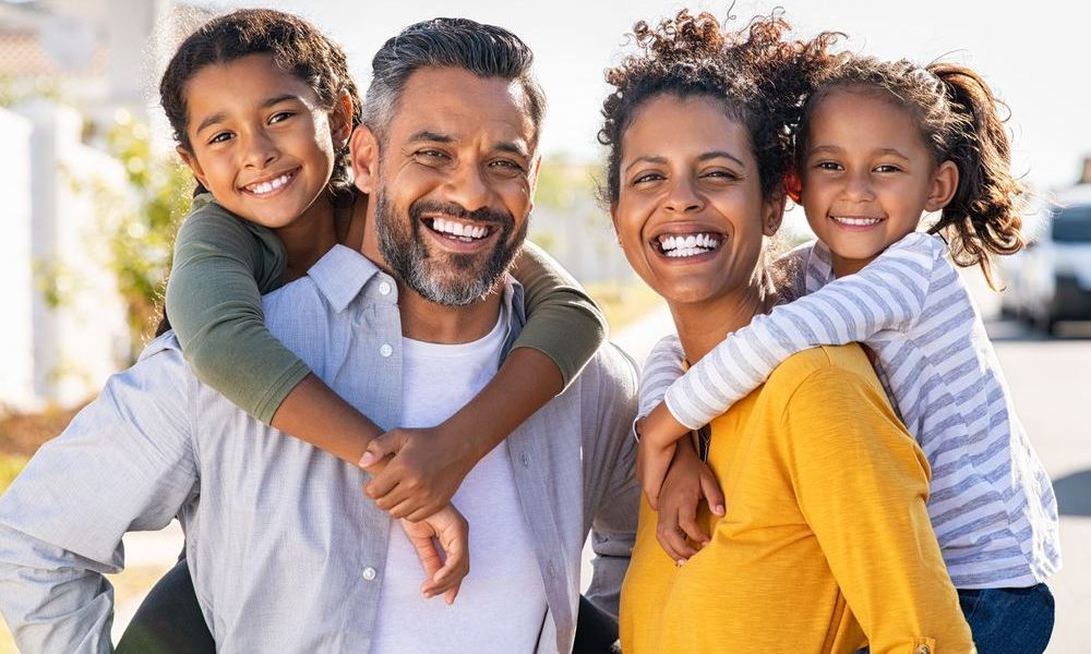 Smiling family with two children outdoors, the parents holding them in bright sunlight.