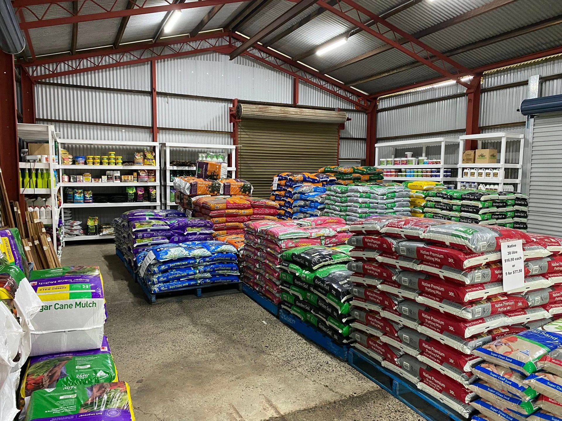 A warehouse aisle lined with metal shelving units filled with various products and stacks of bagged fertilizer on pallets.