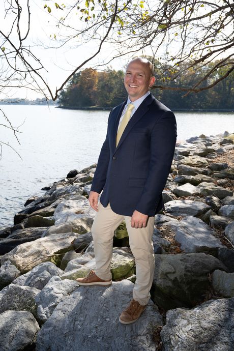 Man standing on rocks by a lake, wearing a navy blazer and khaki pants under trees.