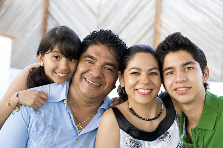 Family of four smiling close together, outdoors with light-colored background.