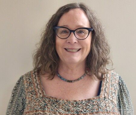 Woman with glasses smiling, wearing patterned top and necklace, against a light wall.