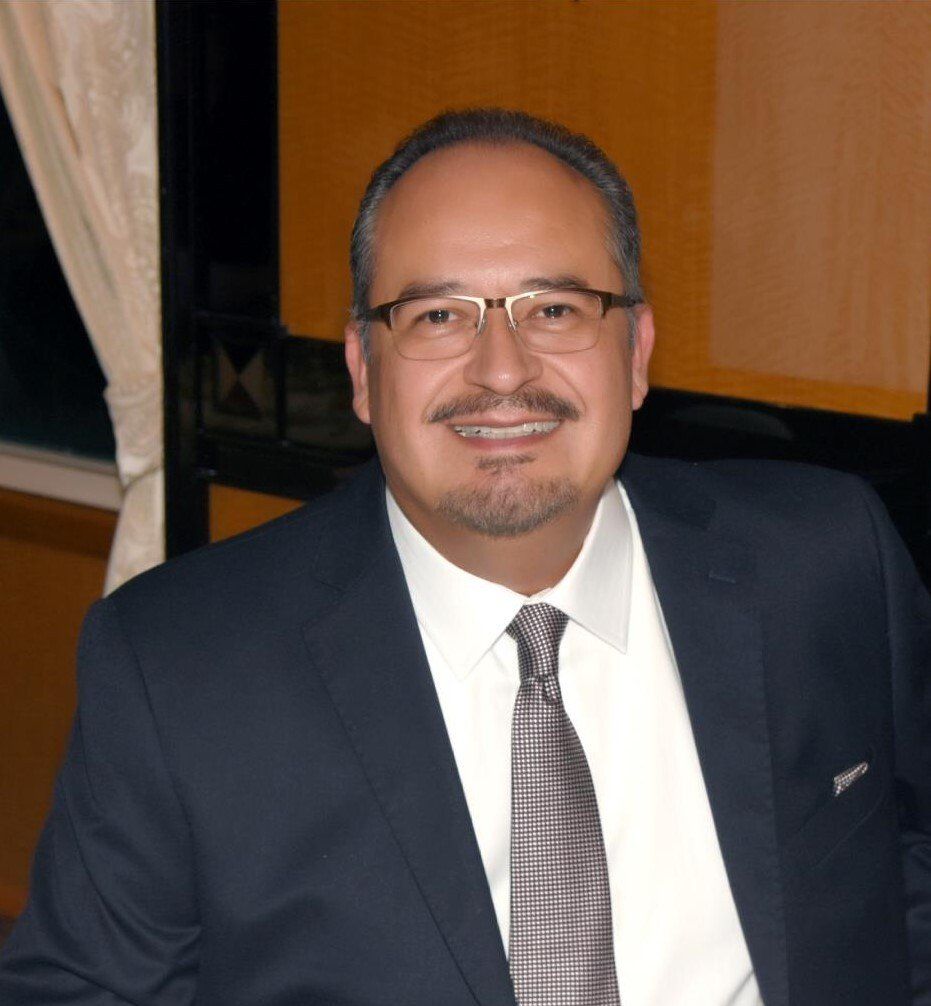 Man wearing glasses and a suit smiles indoors.