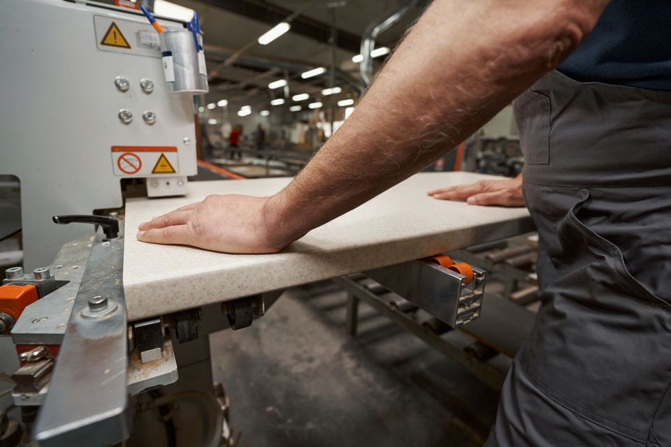 A worker's hands guide a light-colored countertop through a cutting machine in a factory.