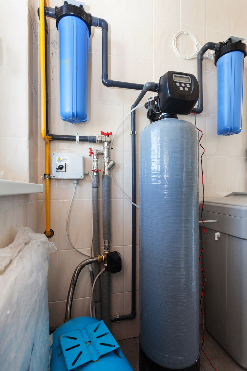 A water filtration system with two blue canisters and a large gray central tank installed against a tiled wall.