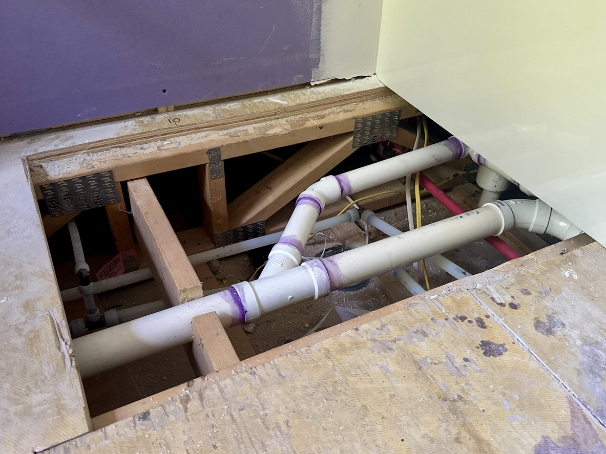 An open floor access area showing white PVC plumbing pipes connected with purple primer against wooden floor joists.
