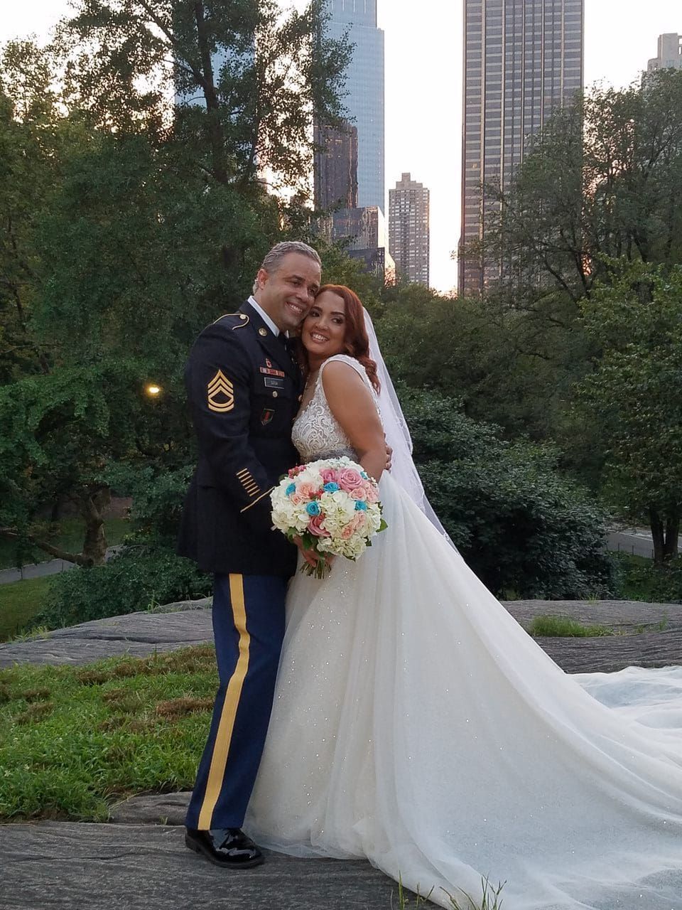 Bride and groom embracing outdoors, bride in white gown holding pastel bouquet, city skyline behind them