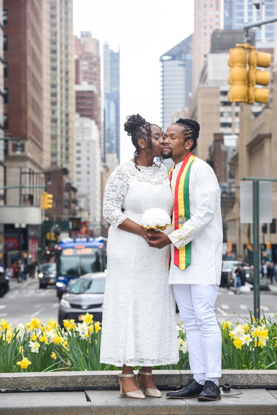 Couple in white outfits holding hands in a city plaza with yellow flowers and tall buildings.