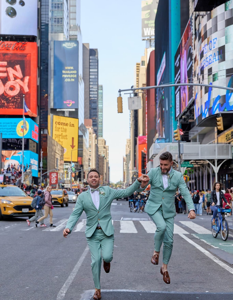 Two men in mint suits jump in Times Square, one holding the other’s hand