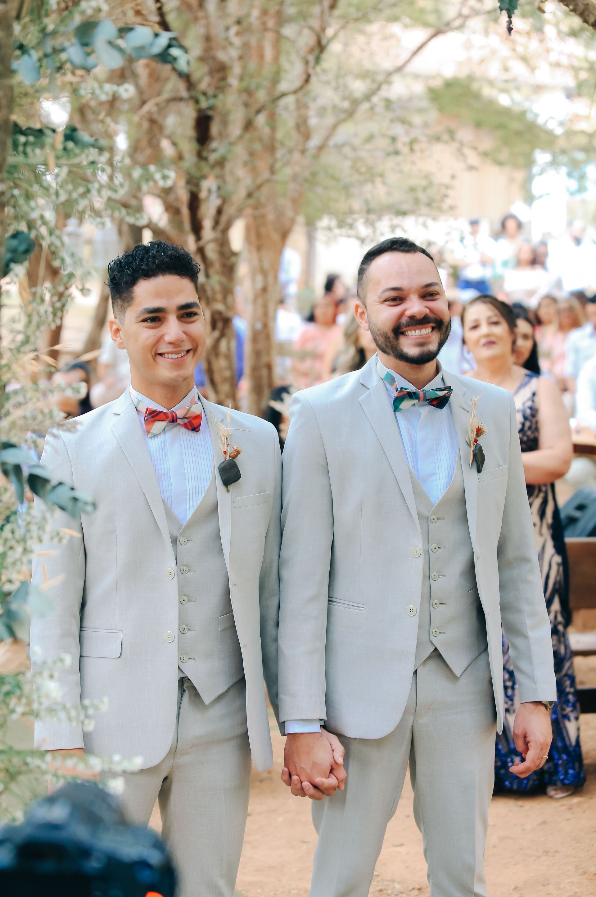 Two grooms in light gray suits smiling and holding hands outdoors