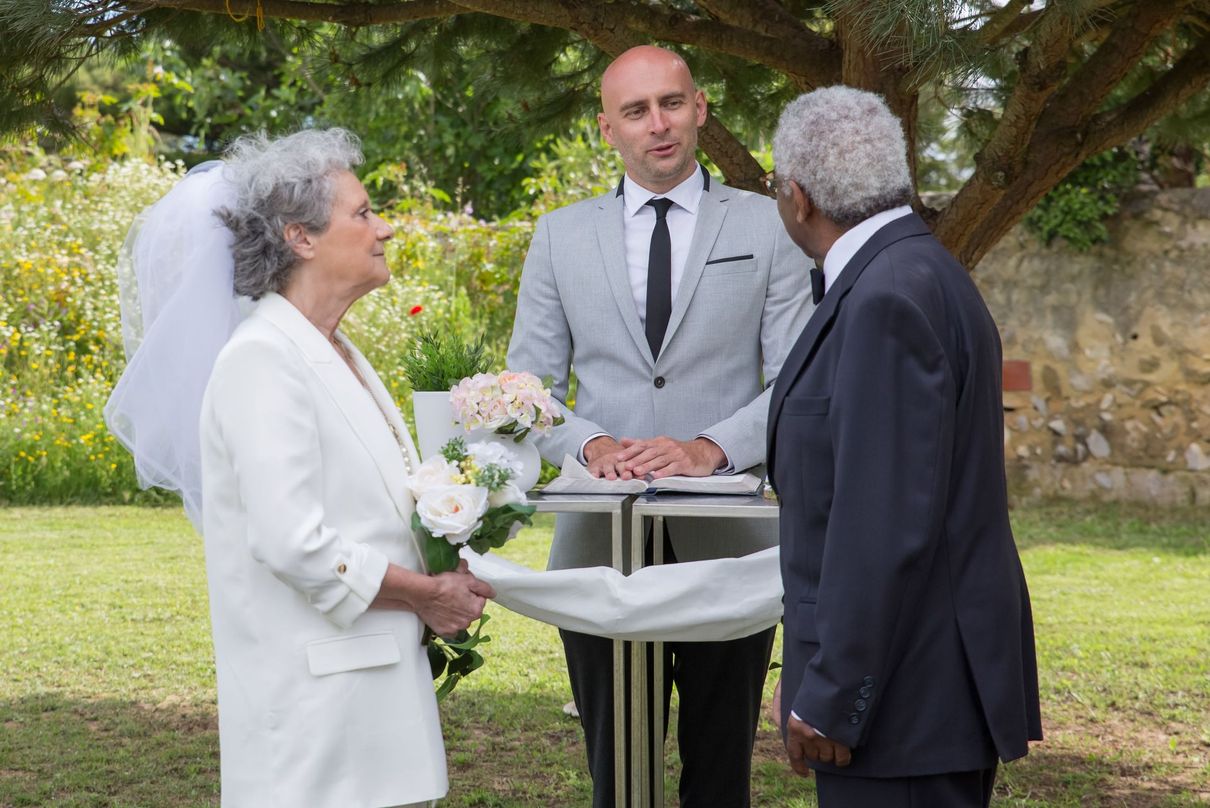 Outdoor wedding ceremony with officiant, bride in white, and groom in dark suit holding hands under a tree