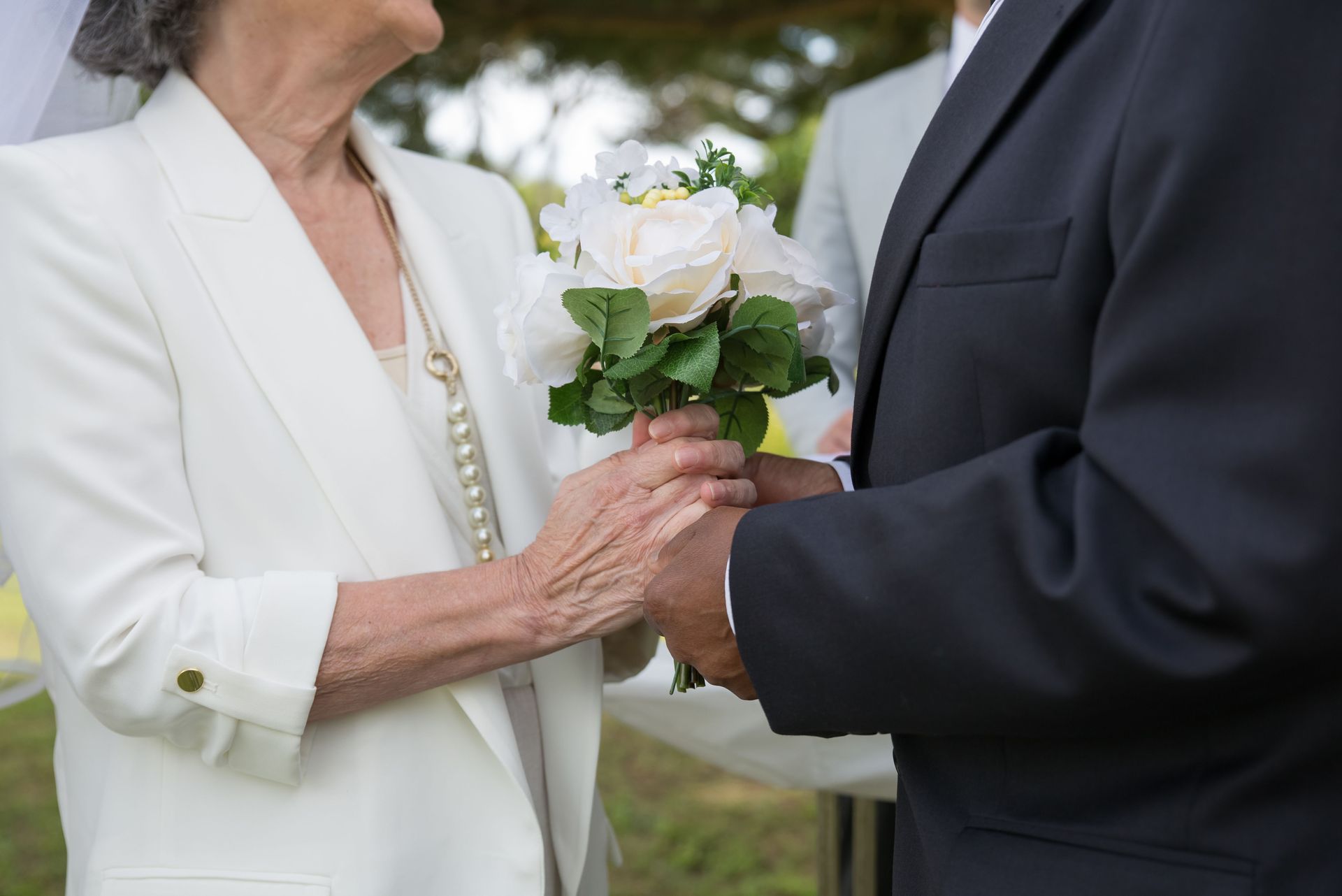Two people in formal attire holding hands and a white flower bouquet outdoors