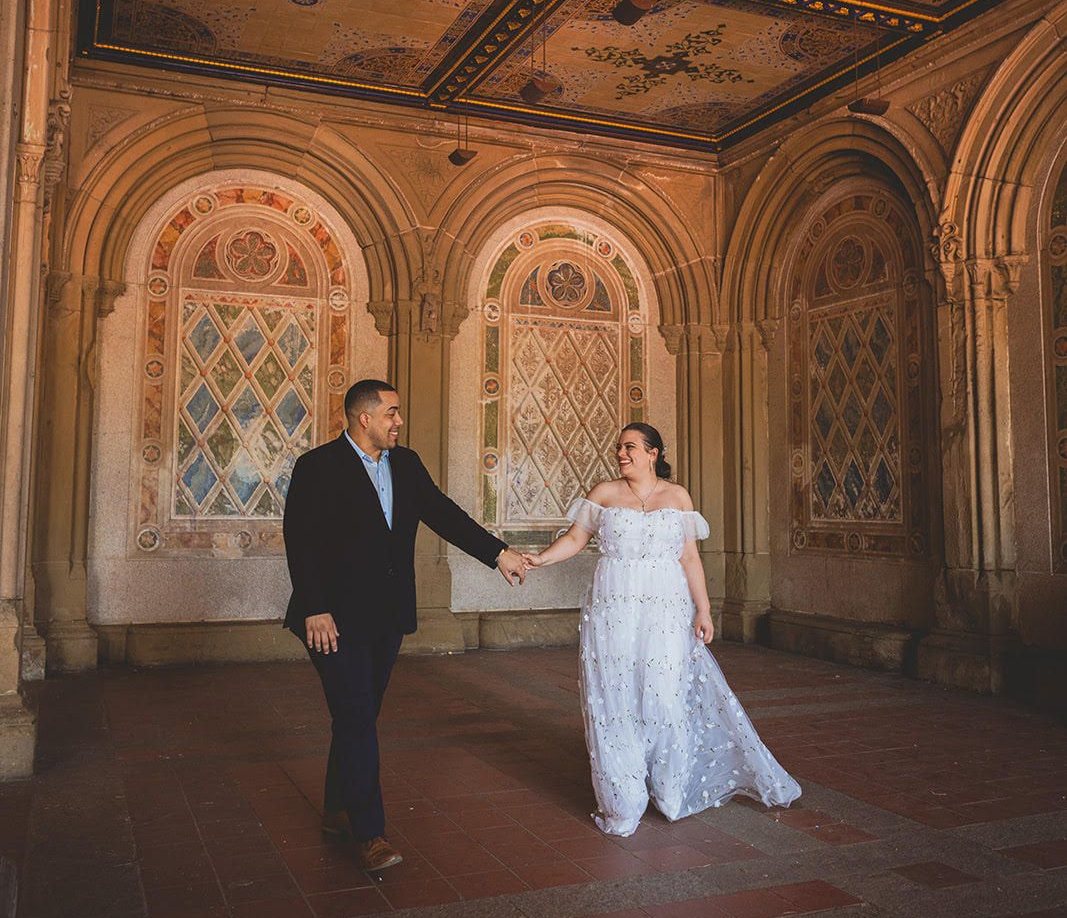 Couple holding hands in an ornate arched room, with the woman in a white dress and the man in a dark suit.