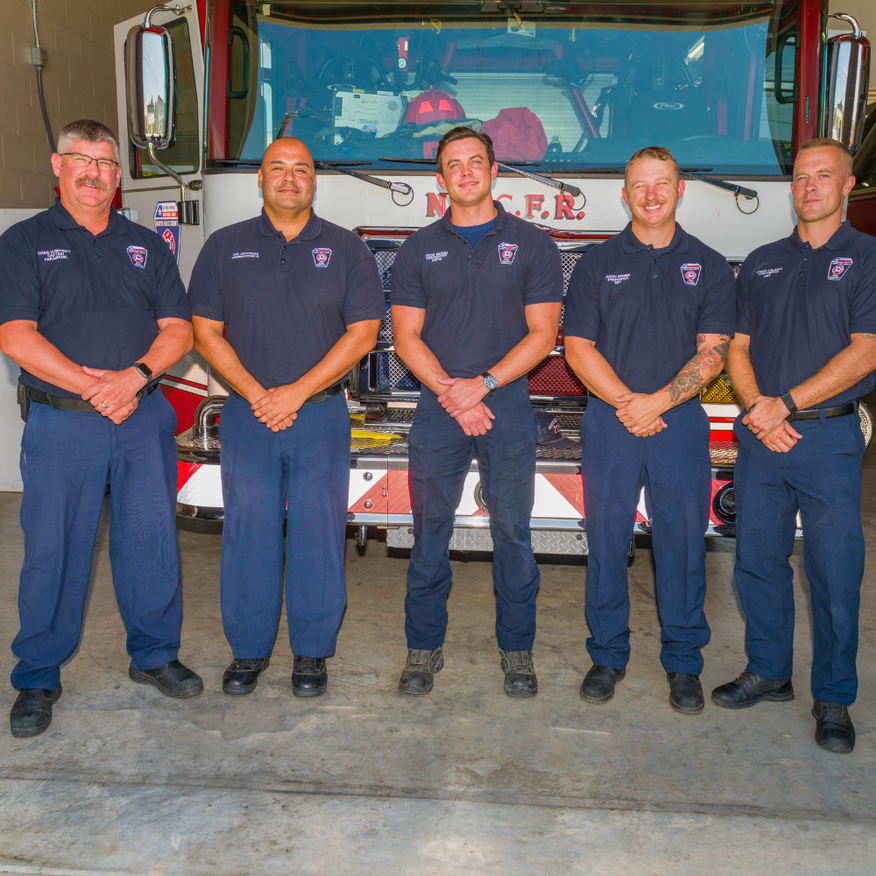 Five firefighters in navy uniforms standing in front of a fire truck in a station garage