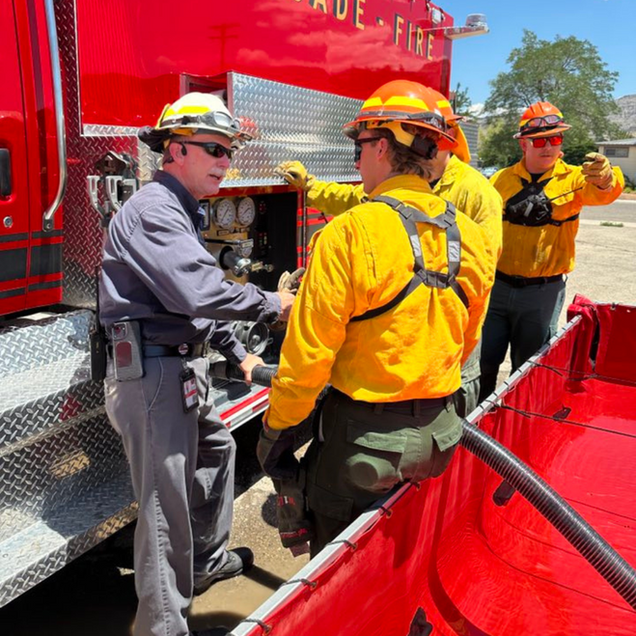 Firefighters in yellow gear talk beside a red fire truck and safety barrier
