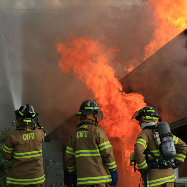 Three firefighters in yellow gear battle a blaze beside a wall of flames and smoke.