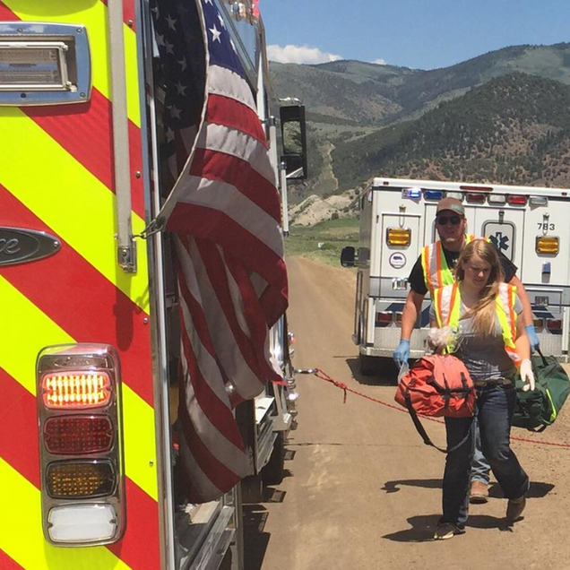 Rescue workers with stretcher beside emergency vehicles on a mountain road, American flag visible.