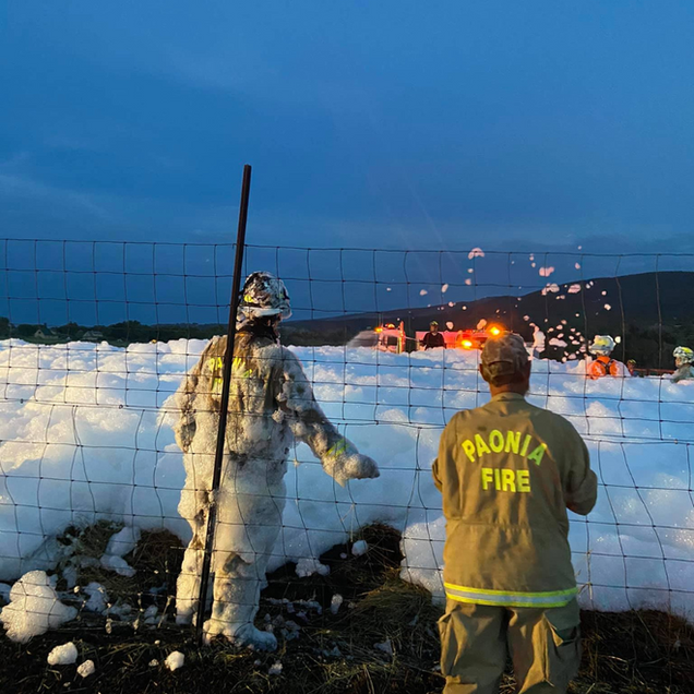 Two firefighters in protective gear monitor flames near a snowy field at dusk.
