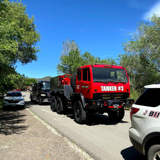 Red tanker truck labeled “TANKER #3” parked on a tree-lined road beside police vehicles
