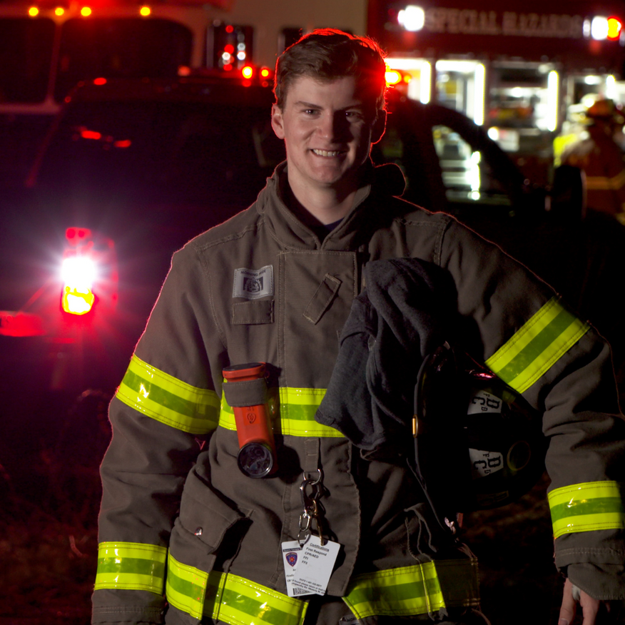 Firefighter in brown turnout gear standing in front of a fire truck at night