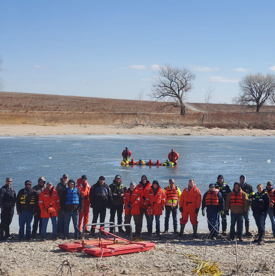 Group of people in orange suits by frozen water, with people in water on rescue devices.